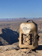 A beige waterproof backpack with black straps and the 'RaCo' logo, set on a rocky desert cliffside with a canyon and blue sky in the background.