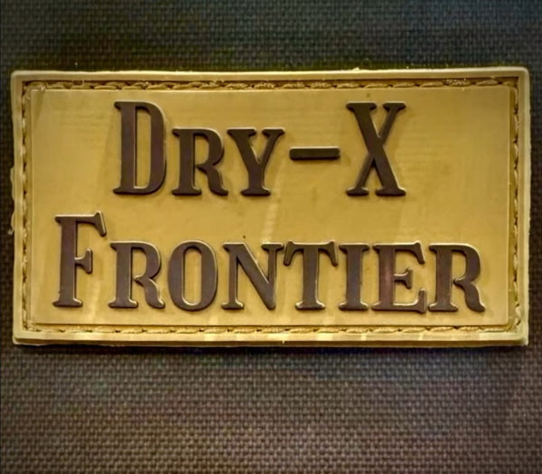 A rectangular gold-toned patch with raised dark lettering reading 'DRY-X FRONTIER' on a dark textured fabric background.