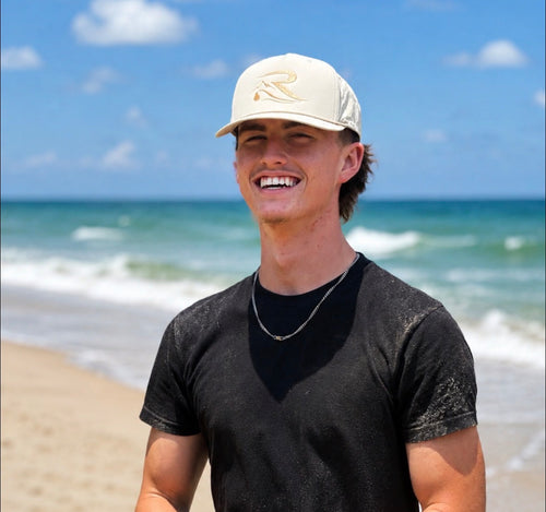 A smiling man standing on a beach wearing a cream trucker cap with a gold embroidered 'R' logo, a black t-shirt, and a silver chain necklace, with the ocean and blue sky in the background.