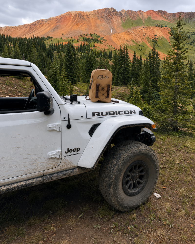 A white Jeep Rubicon off-road vehicle parked on a dirt trail with a mountain and pine forest in the background. A tan canvas bag is hanging from the front hood of the vehicle.